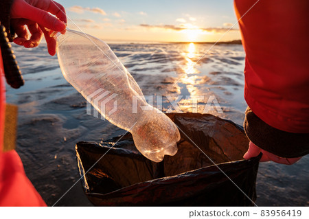 A woman cleans the bank of plastic bottles and puts the trash in a garbage bag. Environmental pollution by plastic. Recyclable packaging. A woman cleans the bank of plastic bottles and puts the trash in a garbage bag. Environmental pollution by plastic. Recyclable packaging. 83956419