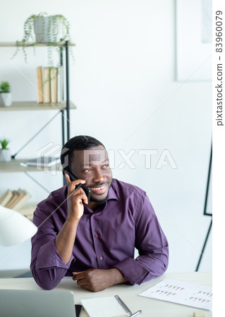 Corporate communication. Happy man. Work break. Smiling guy sitting desk with laptop documents talking smartphone in light room interior. 83960079