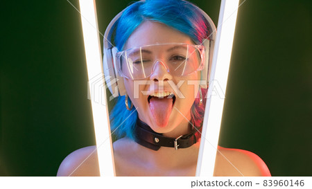 Cyberpunk model. Party girl. Futuristic generation. Funky attractive playful woman sticking tongue out in LED neon light isolated on dark background. Cyberpunk model. Party girl. Futuristic generation. Funky attractive playful woman sticking tongue out in LED neon light isolated on dark background. 83960146