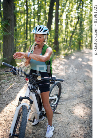 Active senior woman biker sitting on bicycle and setting smartwatch outdoors in forest. 83961325