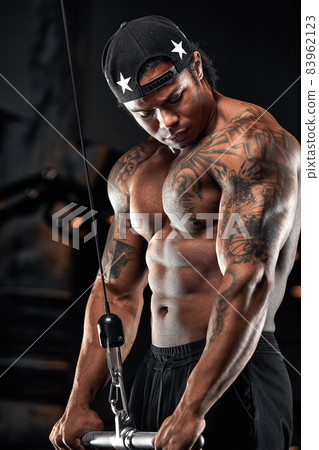 Strong and muscular dark skin man trains on modern equipment in gym. Portrait of muscular pumped up fitness trainer Strong and muscular dark skin man trains on modern equipment in gym. Portrait of muscular pumped up fitness trainer 83962123