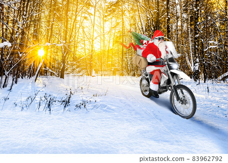 Santa riding a motorbike delivering Christmas or New Year presents, parcels on snow winter landscape at sunset 83962792