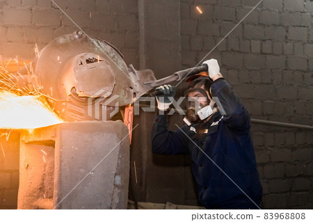 A male worker in a protective helmet, respirator, overalls manages heavy grinding equipment for cast iron concrete tubing with flying sparks in the workshop of an industrial plant A male worker in a protective helmet, respirator, overalls manages heavy grinding equipment for cast iron concrete tubing with flying sparks in the workshop of an industrial plant 83968808
