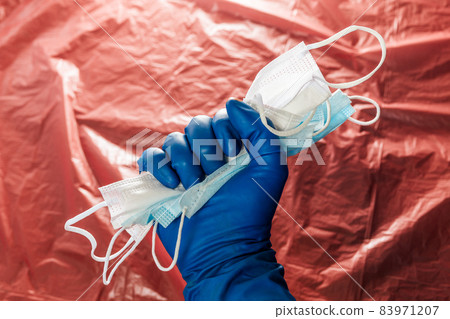 a hand in blue medical glove holding a bunch of face masks on blurry red plastic film background 83971207