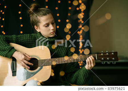 A beautiful teenage girl in Christmas antlers and a green sweater plays the guitar A beautiful teenage girl in Christmas antlers and a green sweater plays the guitar 83975621