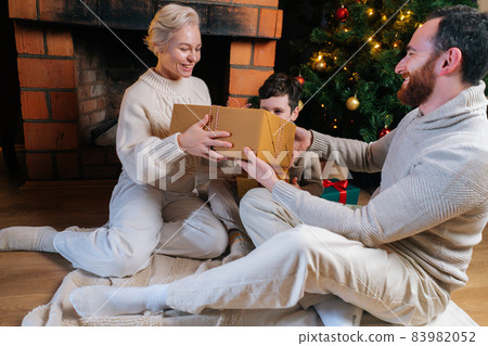 Portrait of happy young woman giving Christmas present from husband and son sitting by fireplace and xmas tree. Portrait of happy young woman giving Christmas present from husband and son sitting by fireplace and xmas tree. 83982052