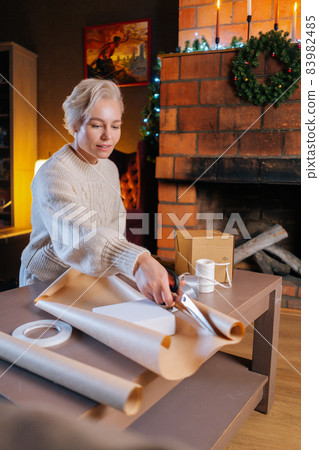 Vertical shot of young woman cutting wrapping craft paper with scissors to wrapping gift boxes of Christmas presents for family 83982485