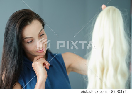 Brunette woman holding blonde wig with long hair and thinking to wear it Brunette woman holding blonde wig with long hair and thinking to wear it 83983563