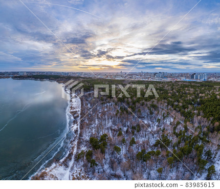 Snow-covered forest on lake shore with the first ice at sunset and the city on horizon, auerial view Snow-covered forest on lake shore with the first ice at sunset and the city on horizon, auerial view 83985615