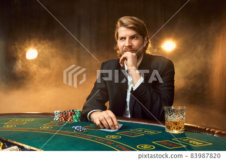 A man in a business suit sitting at the game table. Male player. Passion, cards, chips, alcohol, dice, gambling, casino - it is as male entertainment. A man in a business suit sitting at the game table. Male player. Passion, cards, chips, alcohol, dice, gambling, casino - it is as male entertainment. 83987820