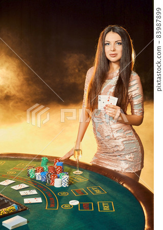 Gorgeous young woman in evening dress standing near poker table with glass of champagne Gorgeous young woman in evening dress standing near poker table with glass of champagne 83987899