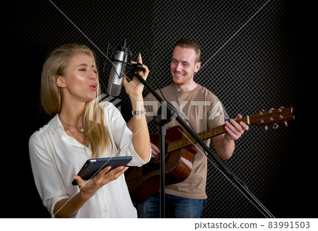 Young female singing with tablet computer in her hand. While the man playing an acoustic guitar in front of black soundproofing walls. Musicians producing music in professional recording studio. 83991503