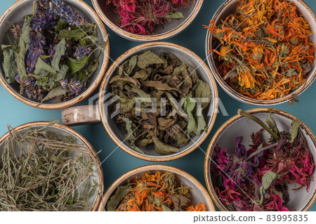 Assortment of dried relaxing tea herbs in colourful cups on blue background close up. Calendula, mint, anise hyssop, monarda didyma, wormwood, sage leaves. 83995835