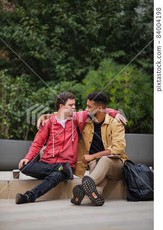 Happy young man with Down syndrome and mentoring friend sitting with arms around outdoors 84004198