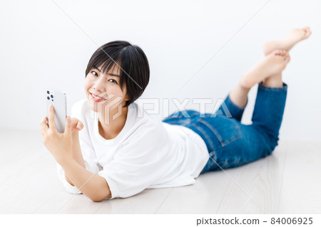 A young woman lying down in the living room and looking at her smartphone 84006925