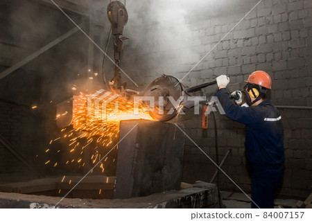 A male worker in a protective helmet, respirator, overalls manages heavy grinding equipment for cast iron concrete tubing with flying sparks in the workshop of an industrial plant 84007157