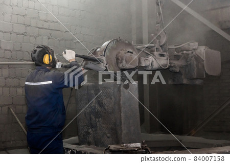 A man in work clothes, and a respirator drives heavy equipment with grinding stone to clean and prepare cast iron concrete tubing in a dirty shop of an industrial plant A man in work clothes, and a respirator drives heavy equipment with grinding stone to clean and prepare cast iron concrete tubing in a dirty shop of an industrial plant 84007158