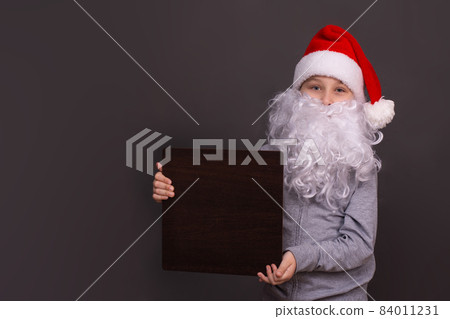 little Santa is holding an empty board for copy space. a boy with a santa beard and a hat holding blank card. on a gray background. little Santa is holding an empty board for copy space. a boy with a santa beard and a hat holding blank card. on a gray background. 84011231