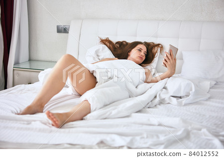 Woman lies in bed and looks at smartphone screen. Woman lies in bed and looks at smartphone screen. 84012552