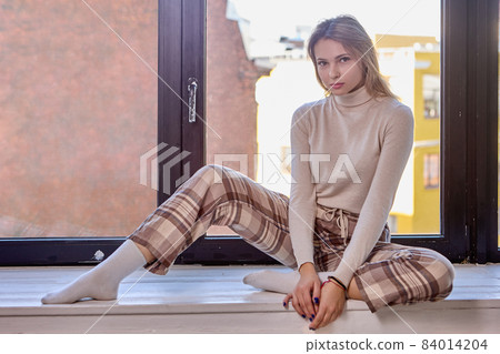 Woman in plaid pants is 24 years old, sitting on windowsill. Woman in plaid pants is 24 years old, sitting on windowsill. 84014204