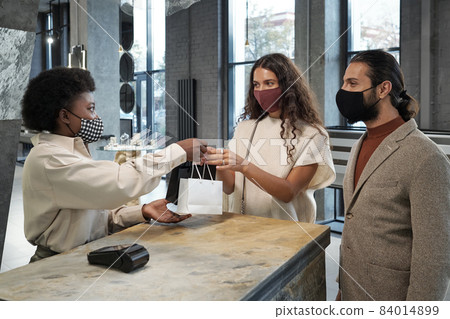 Young woman in protective mask taking paperbags with purchase held by shop assistant over counter 84014899