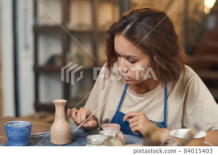 Young woman with paintbrush painting self-made jug or pot 84015403