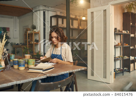 Young ceramist in workwear making notes by workplace 84015476