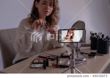 Young caucasian woman is recording an online make-up training video on her mobile. Video blogger girl applies makeup. 84016873