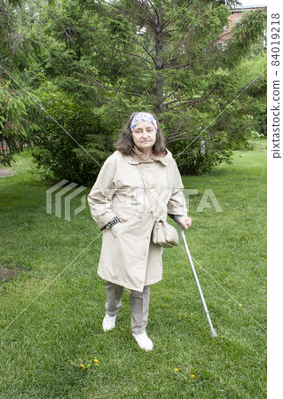 Sad elderly woman with a cane in a jacket walking along the green grass in the park 84019218