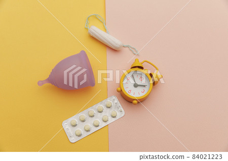 Top view of sanitary cup with pain relief pills, tampon and an alarm clock on colorful background. Menstruation cycle period concept 84021223