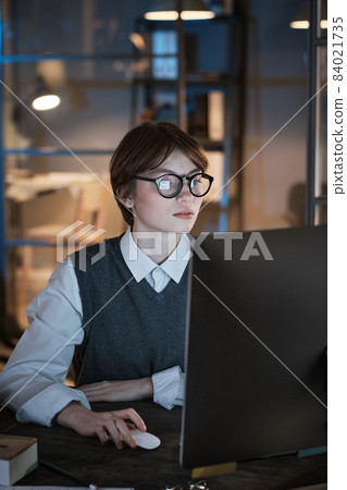 Young businesswoman in eyeglasses sitting in front of the laptop and working on it till late evening at office Young businesswoman in eyeglasses sitting in front of the laptop and working on it till late evening at office 84021735