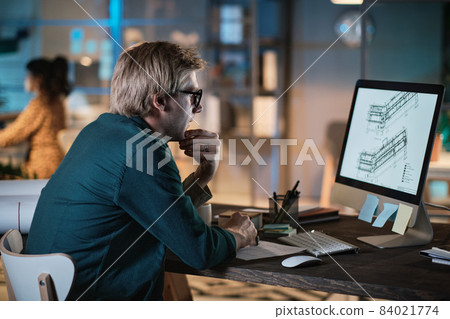 Young designer in eyeglasses sitting at the table and looking at computer monitor, he developing new project at office till late evening 84021774