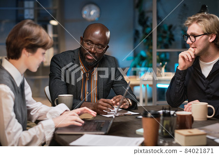Group of business people sitting at the table discussing and using laptop at work during their meeting till late evening at office Group of business people sitting at the table discussing and using laptop at work during their meeting till late evening at office 84021871