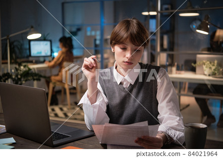 Young businesswoman sitting at the table in front of laptop and examining business documents while working at office till night 84021964