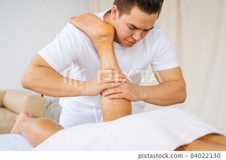 Close-up face of professional male masseur with strong hands massaging calf muscles on lower leg of muscular athlete man after physical sports workout Close-up face of professional male masseur with strong hands massaging calf muscles on lower leg of muscular athlete man after physical sports workout 84022130