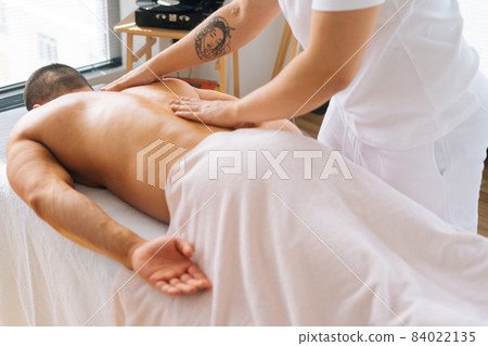 High-angle view of male masseur making gentle stroking movements with strong tattooed hands along spine toward head. High-angle view of male masseur making gentle stroking movements with strong tattooed hands along spine toward head. 84022135