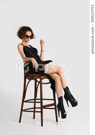 Woman in sunglasses, black dress and pole dance boot sits on tall chair 84023701