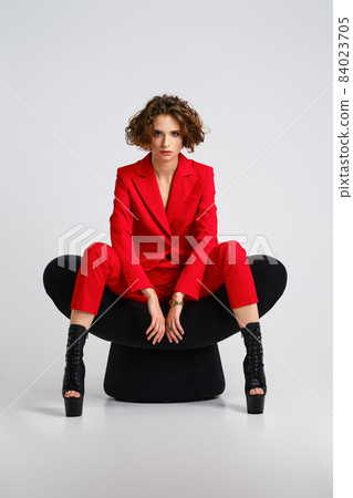 Dancer woman in red pantsui and pole dance boots sits in a soft chair spreading legs 84023705