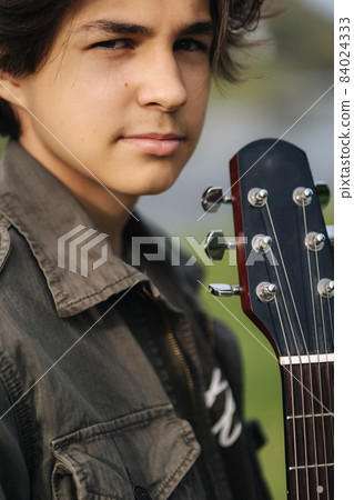 Fashion portrait of long hair boy hold acoustic guitar. Young boy preparing for playing guitar outdoor Fashion portrait of long hair boy hold acoustic guitar. Young boy preparing for playing guitar outdoor 84024333