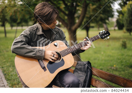 Handsome teenage playing acoustic guitar with capo. Boy sitting on bench and playing music 84024434