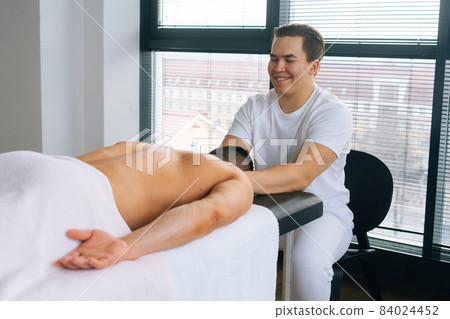 Side view of smiling professional male masseur with strong hands massaging shoulder of muscular sports man lying on stomach at massage table Side view of smiling professional male masseur with strong hands massaging shoulder of muscular sports man lying on stomach at massage table 84024452