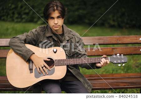 Handsome teenage playing acoustic guitar outdoor in Autumn time. Boy sitting on bench and playing music Handsome teenage playing acoustic guitar outdoor in Autumn time. Boy sitting on bench and playing music 84024518