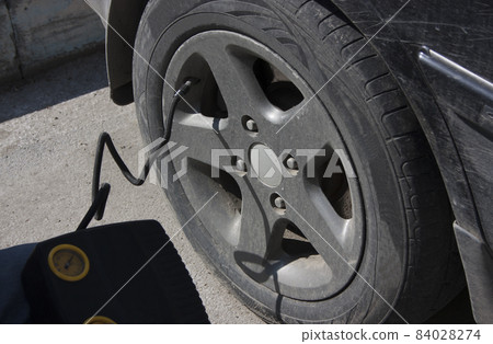 The driver pumps up the wheel of a black car with the help of a compressor The driver pumps up the wheel of a black car with the help of a compressor 84028274