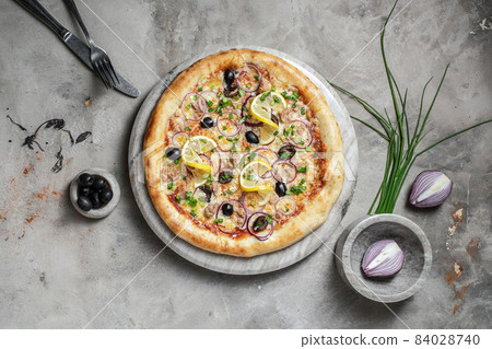 Tasty pizza with  tuna, mozzarella, black olives and onion on marble round tray. Garnished on grey stone backdrop with pizza ingredients 84028740
