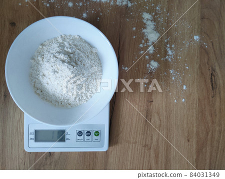 White flour weighing in a bowl on the kitchen scales on wooden table. White flour weighing in a bowl on the kitchen scales on wooden table. 84031349