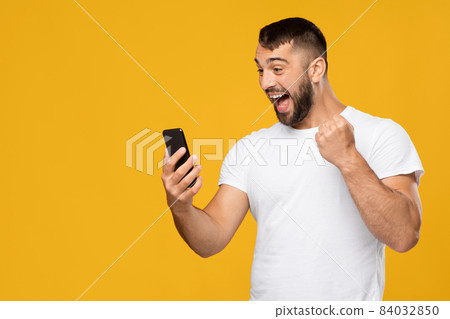 Satisfied excited adult european guy in white t-shirt looking at smartphone and expresses joy, celebrating success 84032850