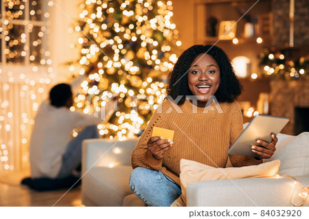Black woman using tablet and credit card, shopping for Xmas gifts online while her boyfriend decorating fir tree at home Black woman using tablet and credit card, shopping for Xmas gifts online while her boyfriend decorating fir tree at home 84032920