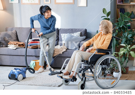 Housemaid using vacuum cleaner to clean the carpet in the room, she helping senior woman who using wheelchair 84033485