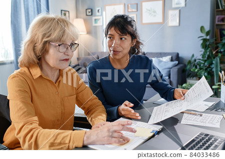 Senior businesswoman in eyeglasses working on laptop with documents at the table together with assistant, they working at home Senior businesswoman in eyeglasses working on laptop with documents at the table together with assistant, they working at home 84033486