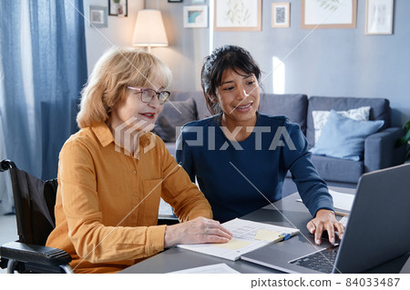 Senior woman in eyeglasses sitting at the table with documents and using laptop together with assistant in the room Senior woman in eyeglasses sitting at the table with documents and using laptop together with assistant in the room 84033487